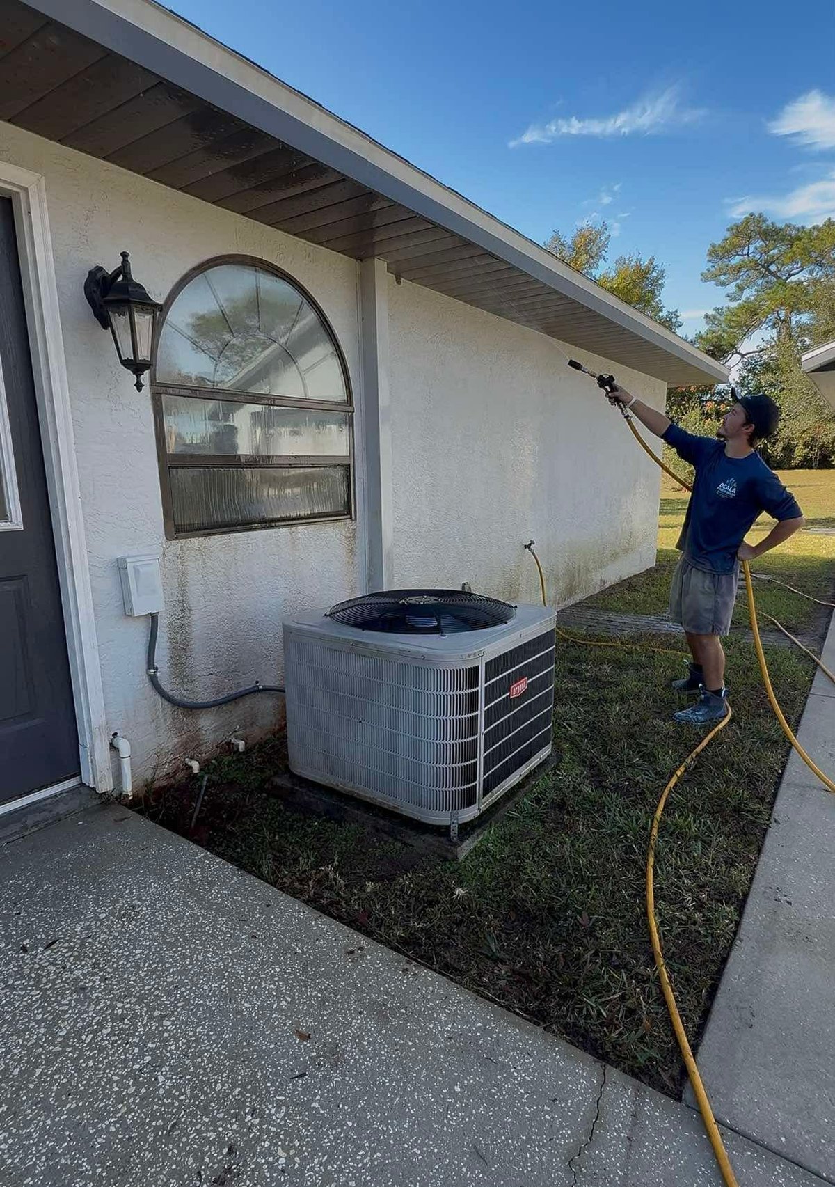 Technician in blue shirt cleaning exterior air conditioning unit with yellow hose at residential home